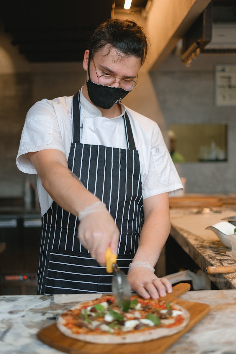 A chef wearing a mask and apron skillfully slices a freshly made pizza in a bustling kitchen.